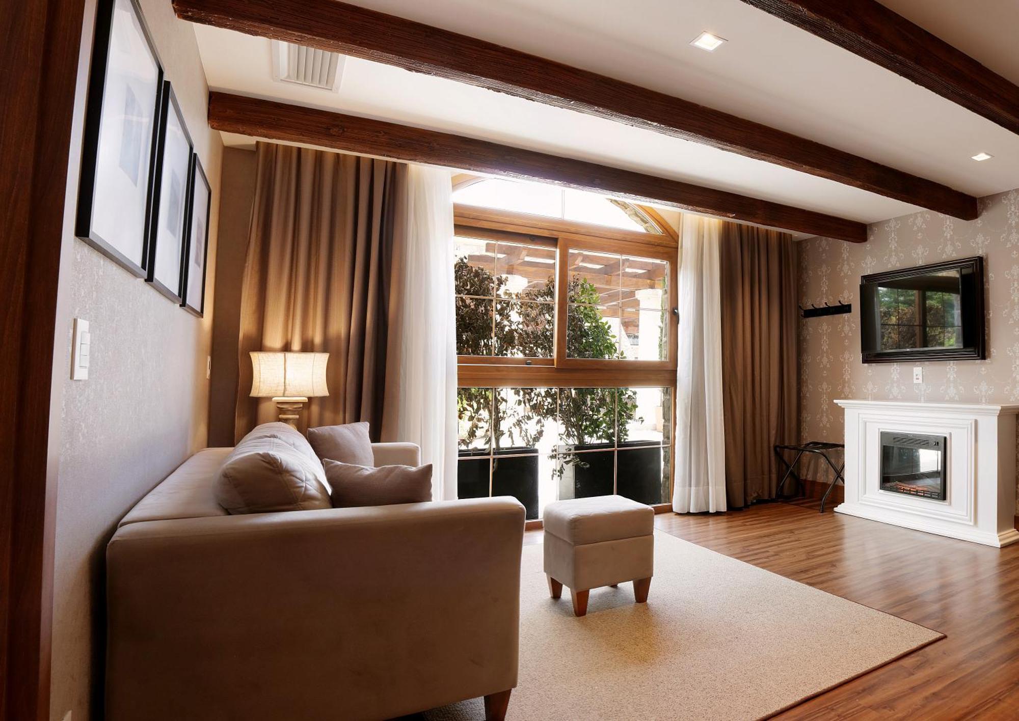 Buona Vitta Gramado Resort & Spa By Gramado Parks Exterior foto The photo depicts a cozy living room setting. It features a light-colored couch, a small ottoman, and a floor lamp beside the couch. Large windows are covered with curtains, allowing natural light to illuminate the space. In the background, there is 